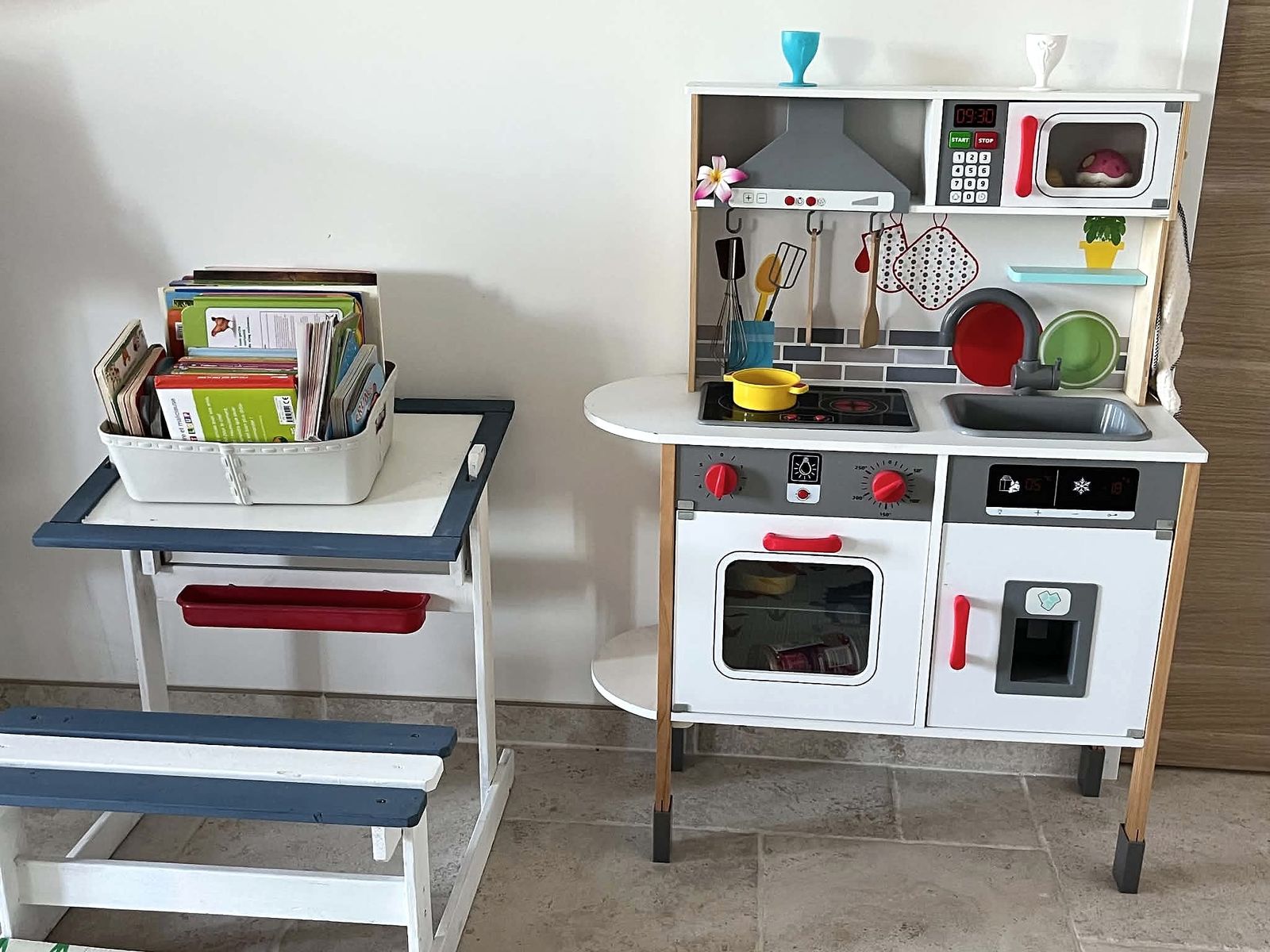 Le coin dînette : une cuisine en bois pour enfants avec ses ustensiles, à côté d’une petite table et son banc où sont rangés des livres pour tout-petits.