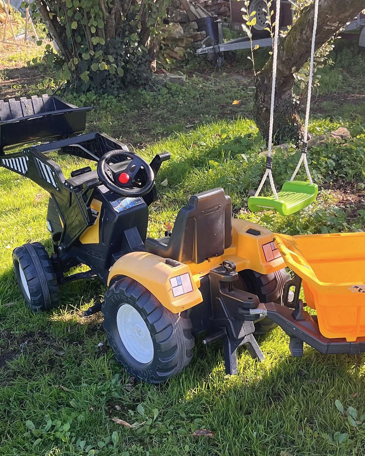 Au jardin, sous un arbre : un tracteur à pédales avec sa remorque, et une balançoire.