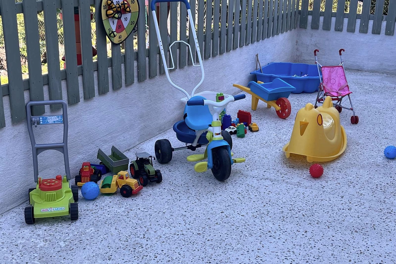 La terrasse de jeux entourée d’une barrière en bois : tricycle, brouette, bac à sable, poussette de poupée, tondeuse et petits véhicules.
